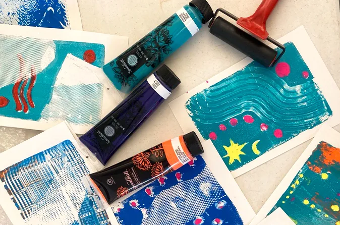 Three tubes of acrylic paint in blue, purple, and orange lie among abstract prints made with a paint roller on white paper.