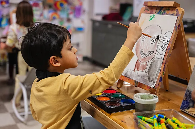 Child in a yellow sweater painting a colorful face on a canvas in the idea Studio.